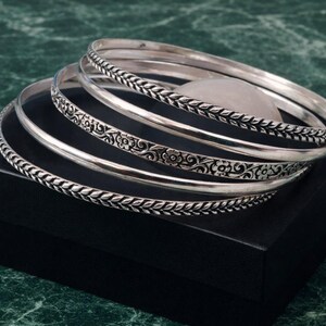 May include: A collection of silver bangles with intricate designs. Some bangles feature a braided pattern, while others showcase floral motifs. The bangles are displayed on a black box against a green marble background.
