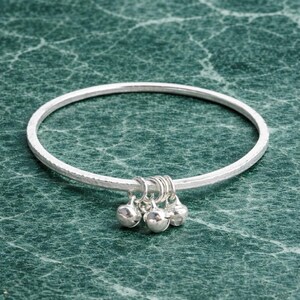 Könnte beinhalten: Ein silbernes Armreif-Armband mit gehämmertem Muster. Das Armband hat vier kleine, runde, baumelnde Anhänger. Der Schmuck wird auf einer strukturierten, grünen Oberfläche präsentiert.