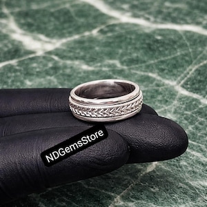 Peut inclure: Une bague en argent avec une bande texturée présentant un motif répétitif. La bague est posée sur un fond sombre, avec une surface semblable à du marbre visible en arrière-plan. La bague a un aspect brillant et métallique.