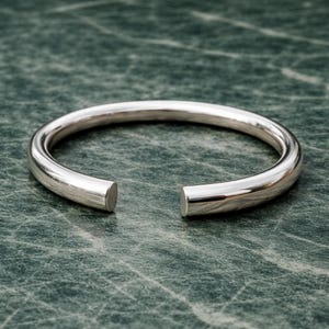 May include: A silver-colored open cuff bracelet. The bracelet has a rounded, tubular design with a gap at the front. The ends are flat and smooth. The bracelet is displayed on a green and gray marbled surface.