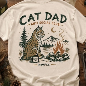 以下が含まれることがあります： クリーム色のTシャツ。オオヤマネコ、キャンプファイヤー、山、そして「CAT DAD ANTI SOCIAL CLUB」の文字が描かれたグラフィックデザイン。オオヤマネコの隣には「NWFC」と書かれたコーヒーマグがあります。デザインは茶色と緑色です。