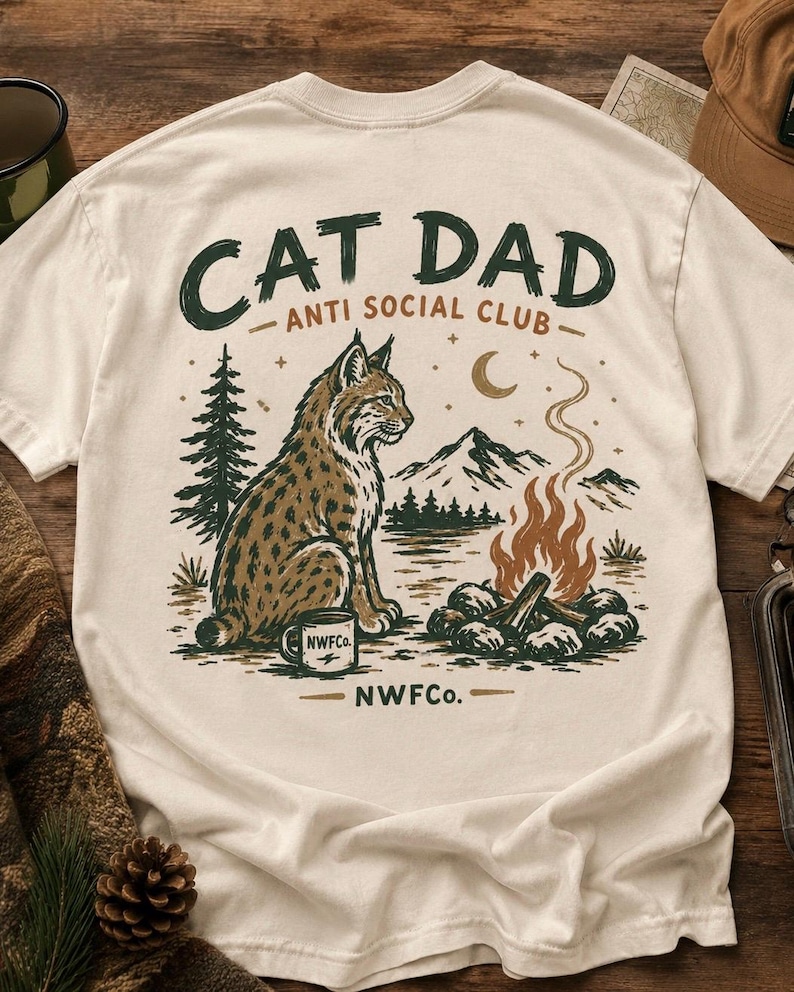 以下が含まれることがあります： クリーム色のTシャツ。オオヤマネコ、キャンプファイヤー、山、そして「CAT DAD ANTI SOCIAL CLUB」の文字が描かれたグラフィックデザイン。オオヤマネコの隣には「NWFC」と書かれたコーヒーマグがあります。デザインは茶色と緑色です。