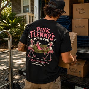May include: A black t-shirt featuring a graphic on the back with three pink flamingos wearing baseball caps and moving a green sofa. The text reads Pink Flemmy's Moving Crew, We lift it. You live it., and NoWhere Fast Co. in pink and white.
