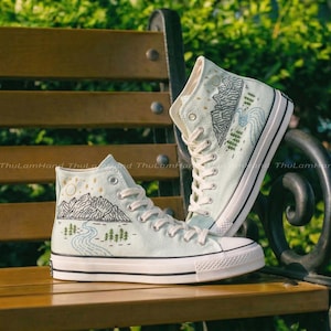 May include: A pair of high-top sneakers with a light blue canvas. The sneakers feature embroidered mountain and river landscape designs in black and blue thread. The soles are white with black trim, and the laces are white.