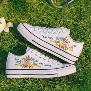 Puede incluir: Zapatillas de lona blancas con bordados florales en tonos rosas, amarillos y azules. Las zapatillas tienen ribetes negros y cordones blancos. Un zapato tiene la fecha "06.26.2026" y el otro "Mrs. Linnard" en azul.