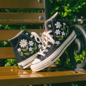 May include: Black high-top sneakers with white embroidered daisy flowers and green leaves. The sneakers have white laces, a white rubber toe cap, and a white sole. The shoes are sitting on a wooden bench.