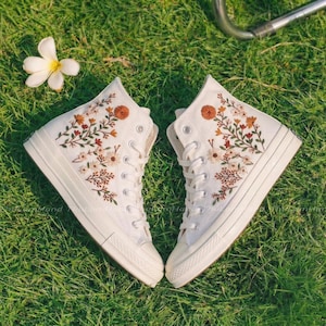 Op de afbeelding: Twee witte hoge sneakers met bloemenborduurwerk in oranje, bruin en crème. De sneakers staan op groen gras, met een witte bloem. De schoenen hebben witte veters en zolen. Botanisch thema.