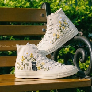 May include: A pair of high-top canvas sneakers in cream color. The sneakers are adorned with embroidered yellow daisies and green leaves. One shoe features a black dog embroidery. The shoes are on a wooden bench.