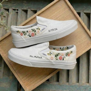 Könnte beinhalten: Ein Paar weiße Slip-on-Sneaker mit gestickten Blumenmustern in Rosa, Gelb und Grün. Ein Schuh zeigt den Text 6/19/26, während der andere den Text Mrs. Matlock aufweist. Beide Schuhe tragen das Vans-Markenlogo an der Seite.