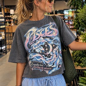 Op de afbeelding: Een antracietgrijs T-shirt met korte mouwen en een afbeelding van twee koivissen die in een cirkel zwemmen, met de tekst PISCES en Empathy level: absorbing the universe. Het shirt wordt gecombineerd met een lichtblauwe broek met witte strepen.