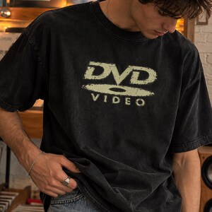 Peut inclure: Un t-shirt noir à manches courtes avec un graphisme délavé et vieilli sur la poitrine, affichant le texte DVD VIDEO dans une police stylisée. Le vêtement présente une encolure ronde et une coupe décontractée en coton noir délavé.