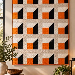 Puede incluir: Un edredón rectangular para colgar en la pared con un patrón geométrico repetitivo de cuadrados naranjas, negros, blancos y gris claro. El edredón está suspendido de una varilla de madera con un cordón fino y muestra una textura acolchada.