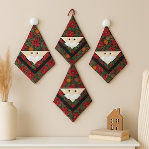 May include: Four Christmas Santa Claus wall hangings. Each is diamond-shaped with a red floral pattern, black trim, and a felt Santa face. The hangings are decorated with orange fruit and white pom-poms. A vase with dried plants and a small house decoration are on a white table.