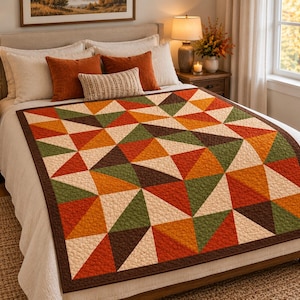 Puede incluir: Una colcha con un patrón geométrico de triángulos en tonos naranja, marrón oscuro, verde oliva y crema. La colcha tiene un borde marrón oscuro y una superficie acolchada con textura, extendida sobre una cama con almohadas a juego.