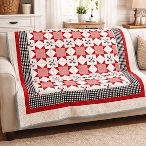 May include: A red, white, and blue quilt on a sofa. The patchwork design includes red squares, white diamonds, and blue floral accents. The quilt is framed by a black and white gingham border.