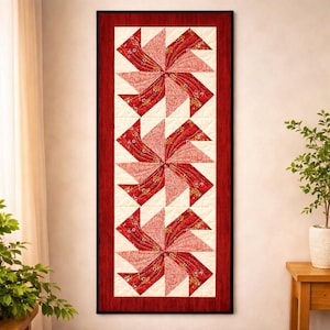 Könnte beinhalten: Ein rechteckiger Wandbehang mit einem Windrad-Design. Der Quilt hat einen roten Rand und eine cremefarbene Mitte. Die Windräder sind aus rot-rosa gemustertem Stoff gefertigt und bilden einen auffälligen Kontrast. Der Quilt ist etwa 30 cm breit und 76 cm lang.