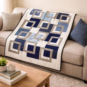 Op de afbeelding: Een gewatteerde sprei met een geometrisch ontwerp in blauw-, grijs- en wittinten is over een beige bank gedrapeerd. De quilt heeft een herhalend patroon van vierkanten en rechthoeken. Er liggen twee sierkussens op de bank en er staat een houten salontafel op de voorgrond.