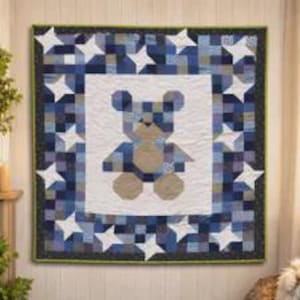Pode incluir: Uma colcha quadrada com um painel central branco apresentando um design de urso de peluche feito com peças em tons de bege e azul. A borda consiste numa grelha de quadrados azuis e brancos com estrelas brancas e um acabamento verde.