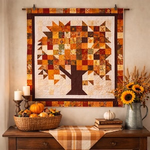 May include: A fall-themed quilt with a tree design hangs above a wooden table. The tree is made of patchwork squares in shades of orange, yellow, and brown. A basket of pumpkins and gourds, sunflowers, and candles decorate the table, creating a cozy autumn scene.