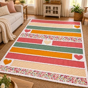 May include: A rectangular area rug featuring horizontal stripes in shades of pink, orange, green, and cream, accented with floral patterns and several solid-colored heart-shaped appliques, all finished with a white frayed-edge border.