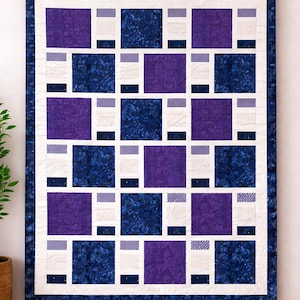 Op de afbeelding: Een gewatteerde wandhanger met een geometrisch patroon. De quilt heeft afwisselende vierkanten in diepblauw en paars, met witte en lichtblauwe accenten. De quilt is omlijst met een donkerblauwe rand en wordt opgehangen aan een houten roede.