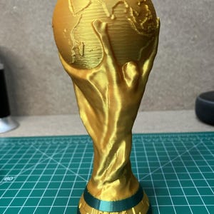 May include: A gold-coloured 3D printed replica of the FIFA World Cup trophy. The object features a spherical globe supported by two stylised human figures, with the text "FIFA WORLD CUP" embossed on the dark green base of the trophy.