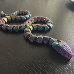 May include: A multi-colored, articulated snake toy. The snake is made of segments that allow it to bend and move. The colors include shades of blue, purple, green, and gold. The snake is lying on a dark surface.