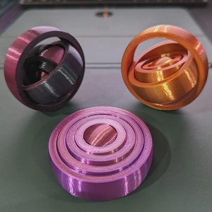 Könnte beinhalten: Drei 3D-gedruckte kinetische Skulpturen in Lila- und Orangetönen. Eine ist eine flache, geschichtete Scheibe. Die anderen beiden sind kugelförmig, mit ineinandergreifenden Ringen und einer griffartigen Struktur. Die Objekte befinden sich auf einer neutralen Oberfläche.