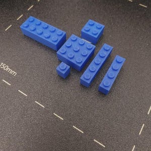 May include: A collection of blue plastic building blocks in various sizes. The blocks include rectangular and square shapes with circular studs on top, designed for interlocking. The blocks are arranged on a dark surface.