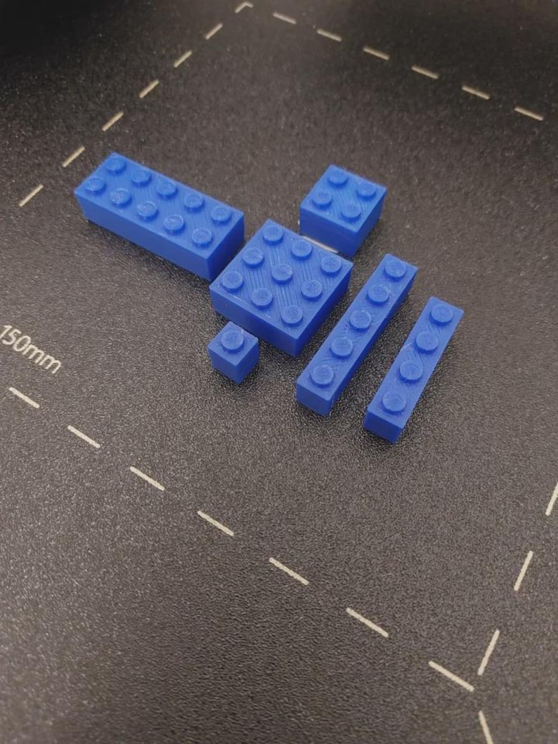 May include: A collection of blue plastic building blocks in various sizes. The blocks include rectangular and square shapes with circular studs on top, designed for interlocking. The blocks are arranged on a dark surface.