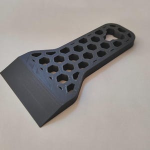 May include: A dark grey scraper with a honeycomb pattern on the handle. The scraper has a flat, angled blade and a handle with hexagonal cutouts. The tool appears to be 3D-printed.