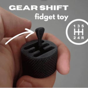 Puede incluir: Un juguete antiestrés con forma de palanca de cambios negra, sostenido en una mano. El juguete tiene una base texturizada y una palanca de cambios. El texto "GEAR SHIFT fidget toy" está encima del juguete.