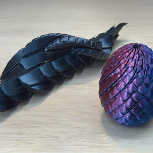 May include: A 3D-printed dragon wing and egg. The wing is black and segmented, with a textured surface. The egg is blue and purple, with a scale-like pattern. The objects are on a light wood surface.