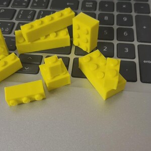 May include: Bright yellow toy building blocks of various shapes and sizes are scattered on a laptop keyboard. The blocks include rectangular and square shapes with protruding studs, suggesting a construction toy theme.