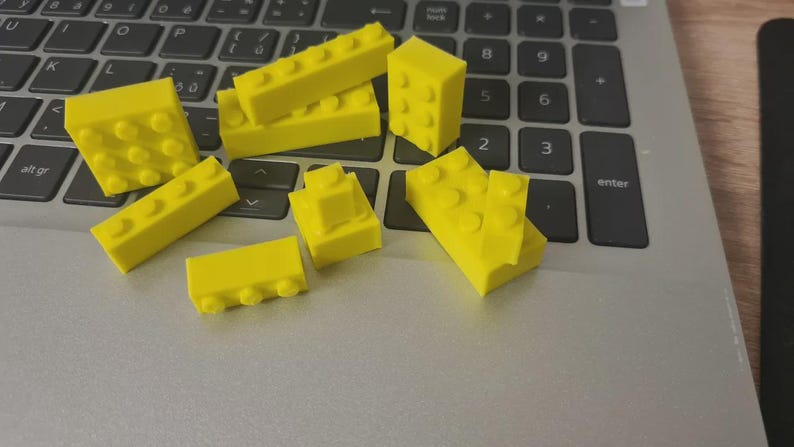 May include: Bright yellow toy building blocks of various shapes and sizes are scattered on a laptop keyboard. The blocks include rectangular and square shapes with protruding studs, suggesting a construction toy theme.