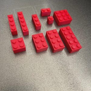 Puede incluir: Una colección de bloques de construcción de plástico rojo de varias formas y tamaños. Los bloques incluyen formas rectangulares y cuadradas con pernos circulares en la parte superior, diseñados para entrelazarse. Los bloques están dispuestos sobre una superficie gris.