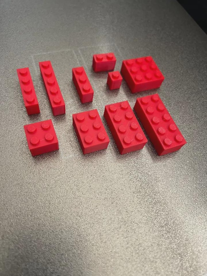 May include: A collection of red plastic building blocks in various shapes and sizes. The blocks include rectangular and square shapes with circular studs on top, designed for interlocking. The blocks are arranged on a gray surface.