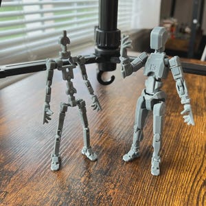 May include: Two grey articulated action figures, designed to mimic human anatomy, stand on a wooden surface. The figures are positioned near a black tripod, suggesting a photography or art context. The figures are poseable, with multiple joints.