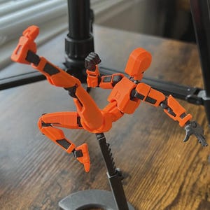 May include: An orange articulated action figure in a dynamic pose, with black accents on the joints. The figure is mounted on a black stand, set against a blurred background with a wooden surface.