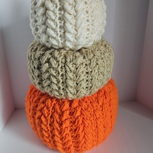 May include: A decorative pumpkin made of three stacked, knitted spheres in cream, tan, and orange. A cinnamon stick serves as the stem. The knitted texture is visible, creating a cozy, autumnal aesthetic. Perfect for fall decor.