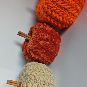 May include: Three handmade crochet pumpkins in varying shades of orange and beige. Each pumpkin has a cinnamon stick stem. The pumpkins are arranged vertically, showcasing their textured surfaces and autumnal theme.