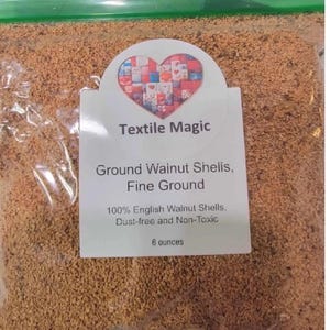 May include: A clear plastic bag filled with fine, brown ground walnut shells. The bag has a white label with the text "Textile Magic" and "Ground Walnut Shells, Fine Ground." The label also states the contents are 100% English Walnut Shells, dust-free, non-toxic, and 6 ounces.