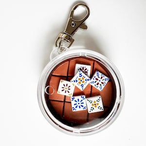 May include: A clear, round keychain with a silver clasp. Inside, a brown square tile design features several small, colorful square tiles with floral patterns in blue, yellow, and red. The keychain is on a white background.