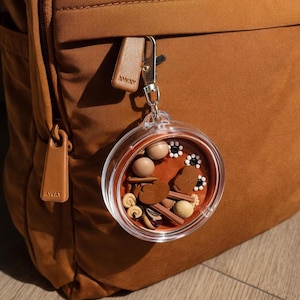 Puede incluir: Un llavero circular transparente con un interior marrón, lleno de elementos decorativos en miniatura. El llavero está sujeto a una mochila marrón con la palabra "AWAY" impresa en el tirador de cuero.