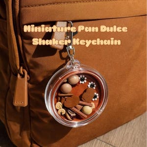 May include: A round, clear plastic shaker keychain containing miniature brown and tan bread-shaped charms and small white flower beads. The keychain features a silver metal clasp and is labeled with the text Miniature Pan Dulce Shaker Keychain.