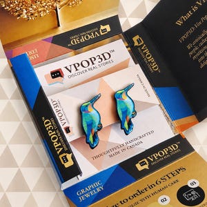 May include: A pair of hummingbird-shaped earrings featuring vibrant blue, green, and yellow patterns. The earrings are displayed on a branded VPOP3D card inside a black and gold presentation box with text including VPOP3D DISCOVER REAL STORIES.