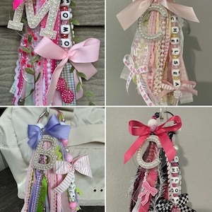 May include: Four colorful handmade keychains with ribbons, beads, and bows. Each keychain features a letter, such as "M", "B", or "C", and a name spelled out with letter beads. The keychains have pink, purple, and white accents.