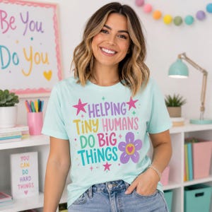 May include: A light mint green short-sleeve t-shirt featuring the colorful text "HELPING TINY HUMANS DO BIG THINGS" in purple, yellow, pink, and teal, accented with pink stars and a purple flower graphic with a yellow center on the front.