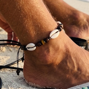 Puede incluir: Pulsera de tobillo con conchas blancas, cuentas marrones y un cordón negro. La pulsera se lleva en un tobillo en una playa de arena con el océano al fondo. Un accesorio con temática playera.