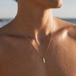 May include: A gold-colored necklace with a rectangular pendant hangs on a person's chest. The chain is delicate, and the pendant is a simple, polished rectangle. The background shows a blurred beach scene with the ocean and sky.